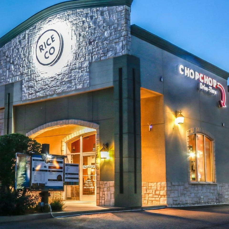 Illuminated Rice & Co. and Chop Chop restaurant building at dusk, featuring stone walls, arched windows, entrance, and drive-through menu.